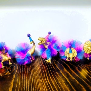 May include: Five gold-colored toy dinosaurs wearing colorful tutus and party hats. The tutus are made of blue, pink, and purple tulle. The dinosaurs are arranged on a dark wooden surface, creating a festive and playful scene.