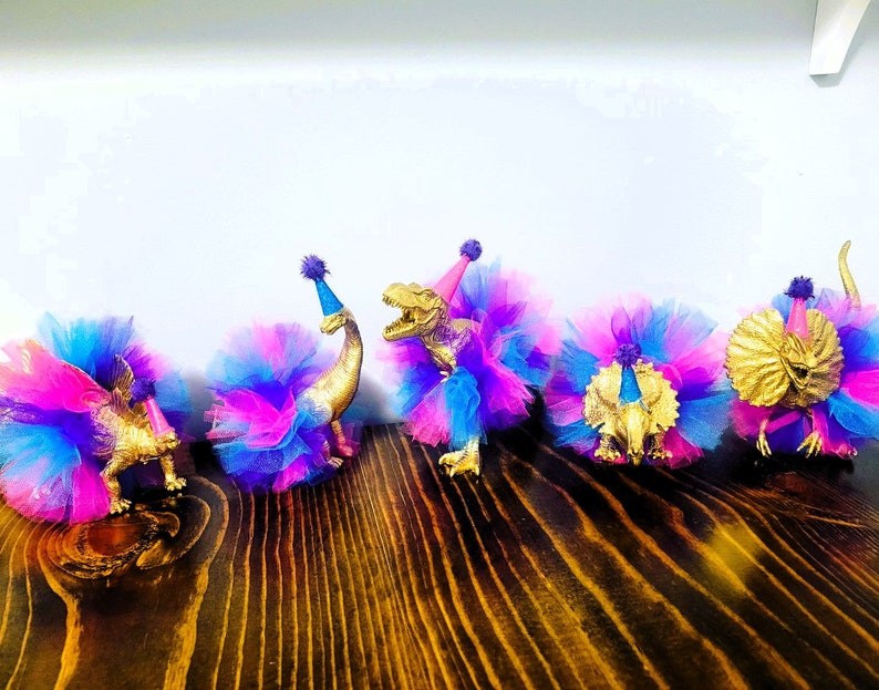 May include: Five gold-colored toy dinosaurs wearing colorful tutus and party hats. The tutus are made of blue, pink, and purple tulle. The dinosaurs are arranged on a dark wooden surface, creating a festive and playful scene.