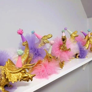 May include: A collection of gold-painted dinosaur figurines, each adorned with a colorful tutu and party hat. The dinosaurs are arranged on a white shelf, creating a whimsical and festive display. The tutus are in shades of pink and purple.