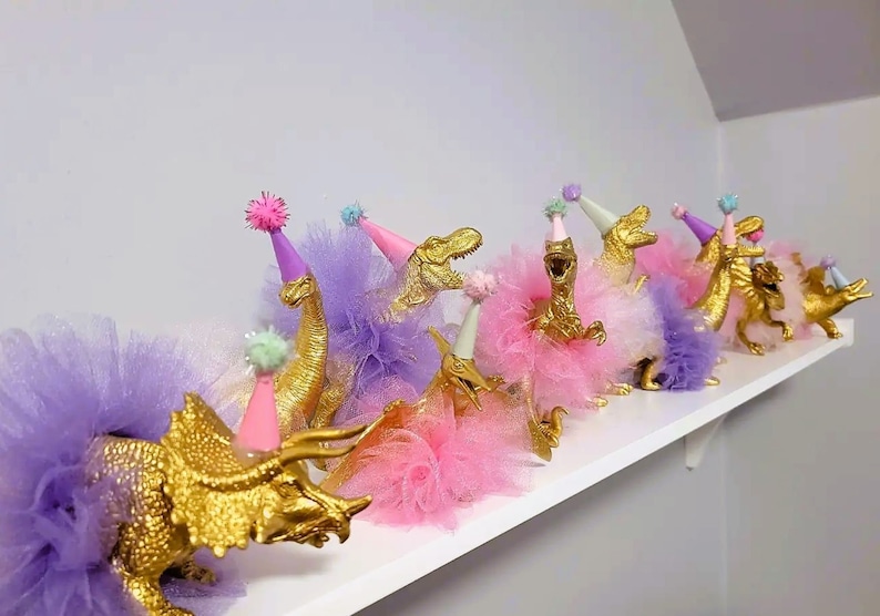 May include: A collection of gold-painted dinosaur figurines, each adorned with a colorful tutu and party hat. The dinosaurs are arranged on a white shelf, creating a whimsical and festive display. The tutus are in shades of pink and purple.