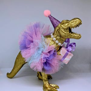 May include: A golden Tyrannosaurus Rex figurine wearing a purple glitter party hat with a pink pom-pom, a multi-colored tutu, and a pearl necklace. The dinosaur holds a small gift box tied with a purple ribbon.