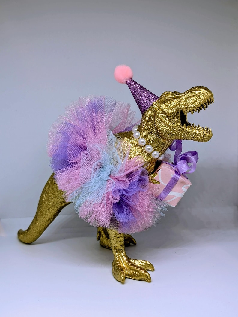 May include: A golden Tyrannosaurus Rex figurine wearing a purple glitter party hat with a pink pom-pom, a multi-colored tutu, and a pearl necklace. The dinosaur holds a small gift box tied with a purple ribbon.