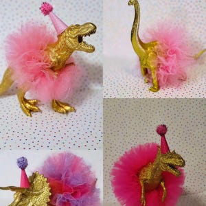 May include: Four gold-colored toy dinosaurs, each adorned with a pink tutu and a party hat. The image includes the text "DINO 1", "DINO 2", "DINO 3", and "DINO 4". The dinosaurs are set against a polka dot background.