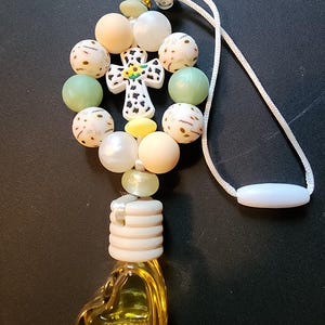 May include: An air freshener featuring a heart-shaped yellow glass bottle and a decorative beaded top. The beads are in various colours, including white, green, and beige, with a cross-shaped charm in the centre. A white cord is attached for hanging.
