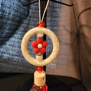 May include: A decorative ornament with a cream-coloured ring adorned with a floral pattern, a red fabric flower, and red and white beads. The ornament has a red, faceted, heart-shaped base and is suspended by a white string.