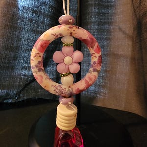 May include: An air freshener with a floral design. The circular top features a pink and white floral pattern. A light purple flower is in the center. The bottom has a clear, red-tinted bottle. The air freshener hangs from a white string.