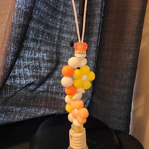 May include: A decorative car air freshener with a clear, diamond-shaped glass bottle. The air freshener features a string of white, orange, and yellow beads and flower-shaped accents. A small orange and clear bead cluster sits at the top.