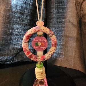 May include: An air freshener with a floral design and the words "I am Worthy I am Enough". The air freshener has a pink and white floral ring, a pink and white beaded top, and a small glass bottle with a pink liquid.