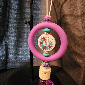May include: A car air freshener featuring a purple ring design with a floral motif. It includes a white cord, a purple and blue bead, and a clear, purple, crystal-like bottom. The air freshener is suspended.