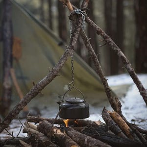 May include: A black metal kettle suspended by a silver chain from a tripod made of three wooden branches. The kettle hangs directly over a small fire built from logs, with visible steam rising from the top lid of the kettle during the heating process.