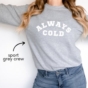 May include: A gray sweatshirt with the text "ALWAYS COLD" printed in white on the front. The sweatshirt is a sport grey crew neck style.