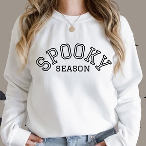 May include: White sweatshirt with the text "SPOOKY SEASON" in black block letters. The text is arched across the chest. The sweatshirt has long sleeves and a crew neck. Black bat silhouettes are in the background.