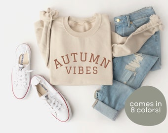 Puede incluir: Una sudadera beige con el texto "AUTUMN VIBES" en letras marrones. La sudadera se combina con jeans azul claro y zapatillas blancas. El texto "comes in 8 colors!" está escrito en un círculo gris.