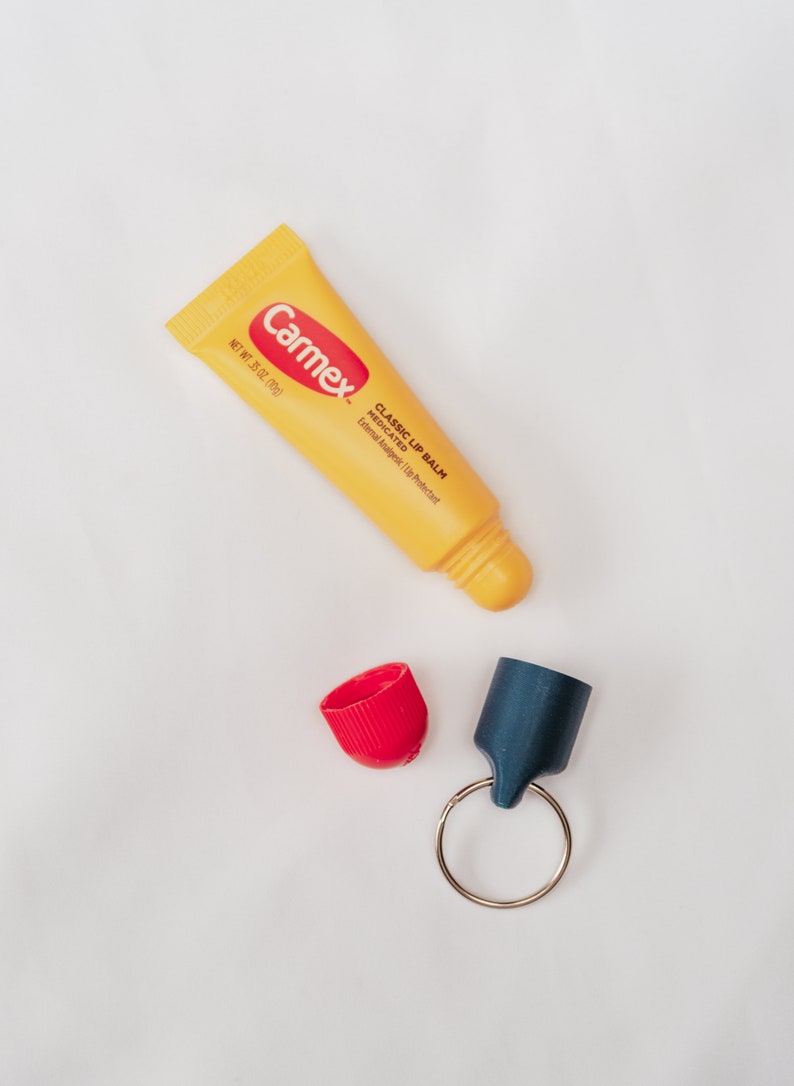 K&ouml;nnte beinhalten: Eine gelbe Tube Carmex Classic Lippenbalsam mit roter Kappe. Die Tube ist mit "Carmex Classic Lip Balm Medicated External Analgesic | Lip Protectant" und "Net Wt. 35 oz. (10g)" beschriftet. Die Tube liegt neben einer roten Kappe und einer blauen Kappe, die an einem silbernen Schl&uuml;sselring befestigt sind.