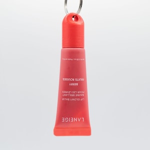 May include: A red tube of Laneige Lip Glowy Balm with a silver key ring attached to the top. The tube is labeled "Lip Glowy Balm", "Baume Brillant", "Pour les Levres", "Berry", and "Fruits Rouges".