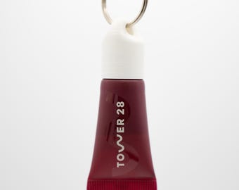 May include: A small, maroon-colored tube of lip gloss with the text "TOWER 28" printed vertically in white. The tube has a white cap and a silver keyring attached. The product is against a white background.