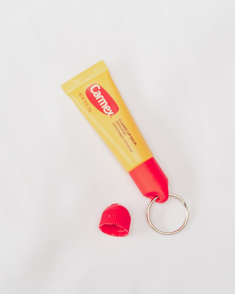 K&ouml;nnte beinhalten: Eine gelbe Tube Carmex Classic Lippenbalsam mit roter Kappe und einem silbernen Schl&uuml;sselring. Die Tube ist mit "Classic Lip Balm Medicated" und "External Analgesic/Lip Protectant" beschriftet.