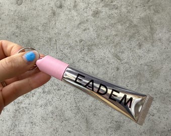 Könnte beinhalten: Eine silberne Tube mit dem schwarzen Aufdruck "EADEM". Sie hat eine rosa Kappe und einen silbernen Schlüsselring. Das Produkt wird von einer Hand mit blau lackiertem Nagel gehalten.