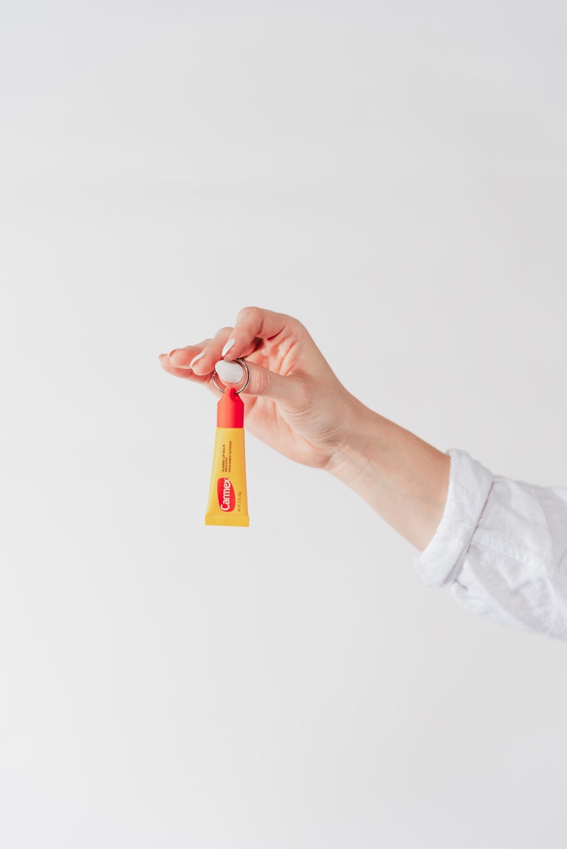 K&ouml;nnte beinhalten: Eine Hand h&auml;lt eine kleine gelbe Tube Carmex Lippenbalsam, die an einem silbernen Schl&uuml;sselring befestigt ist. Der Lippenbalsam ist mit "Carmex" und "Lippenbalsam mit Sonnenschutz SPF 15" beschriftet.