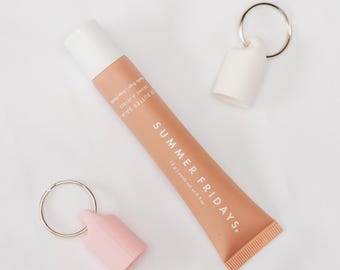 May include: A tube of Summer Fridays lip butter balm in a peach color, with the text "SUMMER FRIDAYS" and "BUTTER BALM" printed on it. Two keychains with white and pink cases are also in the image.