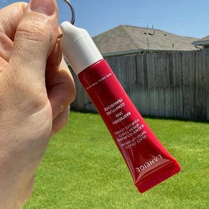 May include: A red and white tube of Laneige Glaze Craze Tinted Lip Serum in Raspberry Jam. The tube is held by a silver keyring. The text on the tube includes "Raspberry Jam" and "Tinted Lip Serum". The background is a green lawn and a blue sky.