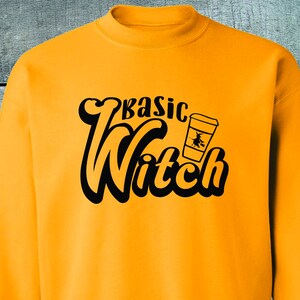 Könnte beinhalten: Senfgelbes Sweatshirt mit dem Schriftzug "Basic Witch" in einem Retro-Schriftstil. Eine schwarze Grafik einer Hexe auf einem Besen in einer Kaffeetasse befindet sich neben dem Text. Das Sweatshirt ist aus weichem Material.