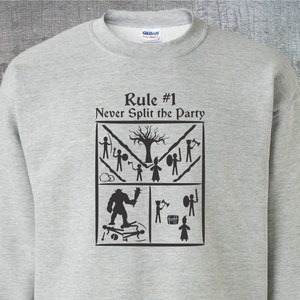 Könnte beinhalten: Ein graues Sweatshirt mit einem schwarzen Grafikdruck, auf dem "Regel Nr. 1 Teile niemals die Gruppe auf" steht. Die Grafik ist ein Quadrat, das in vier Abschnitte unterteilt ist, die jeweils ein anderes Szenario aus einem Rollenspiel darstellen.