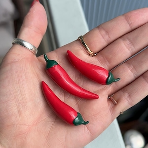 May include: Three small, red chili pepper-shaped objects with green stems. The peppers are smooth and glossy, and appear to be handmade. The peppers are resting on a light-colored surface.