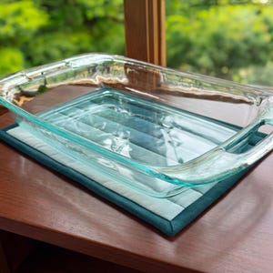 May include: A clear glass baking dish with handles rests on a quilted teal and light blue pot holder. The pot holder is square with a dark teal border. The baking dish is empty and sits on a wooden surface.