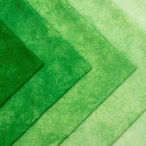 May include: An assortment of green fabric swatches in varying shades, from deep emerald to pale lime. The fabric has a textured, mottled appearance, suggesting a batik or hand-dyed technique. The arrangement creates a gradient effect.