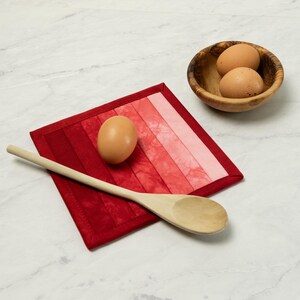 May include: A red and pink ombre quilted hot pad with a brown egg and a wooden spoon. Two brown eggs sit in a small wooden bowl. The items are arranged on a white marble surface.