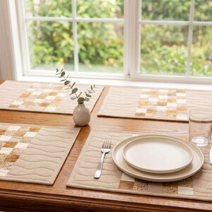May include: A set of rectangular fabric placemats featuring a central panel of quilted patchwork squares in shades of tan, cream, and gold, bordered by solid tan fabric with wavy stitched lines. The placemats are arranged on a wooden table.