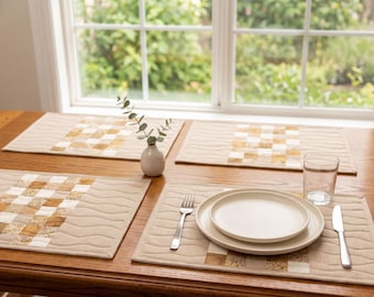 May include: Four rectangular fabric placemats featuring a central patchwork strip of tan, brown, and white squares flanked by solid tan panels with wavy quilted stitching. One placemat holds a white plate, a fork, a knife, and a glass.