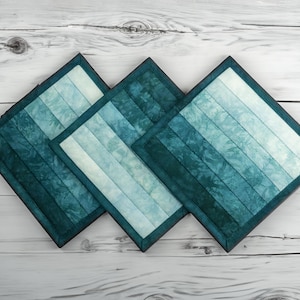 May include: Three square quilted coasters in shades of teal and blue. Each coaster features a gradient design with horizontal stripes and a dark teal border. The coasters are arranged on a light wood surface.