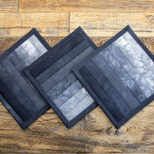 May include: Three square fabric pot holders featuring a gradient design that transitions from solid black to mottled shades of gray and white. Each piece is constructed with parallel quilted strips and finished with a dark fabric border.