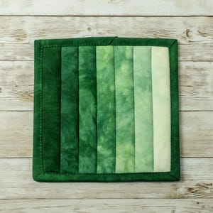 May include: A green quilted pot holder with a gradient of green shades. The pot holder is made of fabric and has a stitched border.