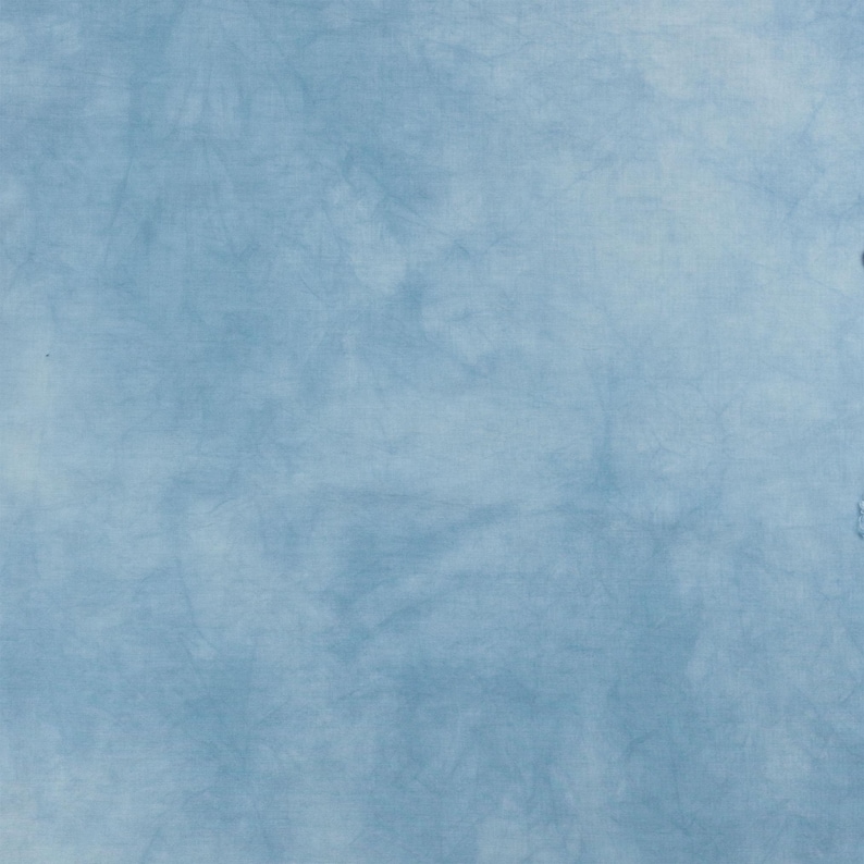 K&ouml;nnte beinhalten: Ein hellblauer Stoff mit einem subtilen, gesprenkelten Batikmuster. Die Oberfl&auml;che weist weiche, wolkenartige Variationen in der Farbintensit&auml;t auf, die von blassem Himmelblau bis zu etwas tieferen T&ouml;nen reichen und eine texturierte Optik erzeugen.