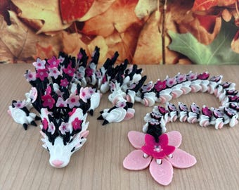 May include: An articulated dragon figurine featuring a white, black, and pink colour scheme. The dragon is adorned with numerous small pink and white flower blossoms along its back and head, ending in a large pink flower tail piece.