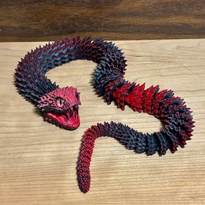 Peut inclure: Une figurine de serpent articulée et flexible dotée d'un corps segmenté aux écailles pointues. Le serpent présente un dégradé de noir profond et de rouge vif, avec une gueule grande ouverte révélant des dents et un œil à pupille verticale.