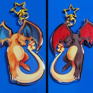 May include: Two acrylic keychains featuring a Charizard design. One keychain has an orange Charizard with blue wings, the other has a black Charizard with red wings. Both keychains have a gold star charm and a gold chain.