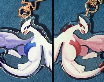 Puede incluir: Dos llaveros de acrílico con una criatura estilizada similar a un dragón. Un llavero muestra un cuerpo blanco con alas azules y detalles en azul oscuro. El otro llavero muestra un cuerpo blanco con alas rosas y detalles en color rojo.