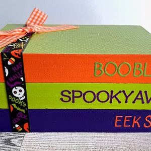 May include: A three-tiered wooden box with a green, orange, and purple paint scheme. The top tier is green with white polka dots and says "BOO BLVD" in orange letters. The middle tier is orange and says "SPOOKY AVE" in purple letters. The bottom tier is purple and says "EEK ST" in orange letters. The box is decorated with a black and white ribbon with a Halloween theme.