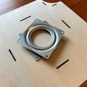 May include: Two silver metal swivel plates for furniture, stacked on top of each other, on a light brown wooden surface.