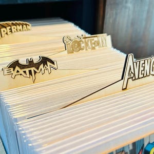 May include: A close-up of a wooden comic book storage shelf with dividers labeled "Superman", "Batman", "Lockey", "The Avengers", and "Superman".