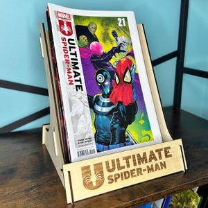 Puede incluir: Un expositor de cómic de madera que contiene un número de Ultimate Spider-Man. El cómic presenta una colorida ilustración de Spider-Man y otros personajes. El expositor tiene las palabras "ULTIMATE SPIDER-MAN" grabadas en la parte delantera.