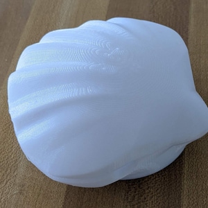 May include: A white, 3D-printed seashell object with a ribbed, fan-like texture across its surface. The object features a rounded, clam-like shape with visible horizontal layer lines from the printing process, creating a matte finish.