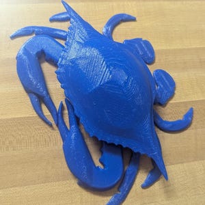 May include: A three-dimensional blue crab figurine featuring a textured surface with visible horizontal print lines. The crab is depicted with its claws extended and legs positioned outward, showcasing a detailed shell structure and sharp edges.
