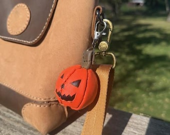 May include: A brown and tan leather handbag with a small orange pumpkin charm. The pumpkin charm has a carved face with black details. The bag has a handle and a strap with a gold-colored clasp.