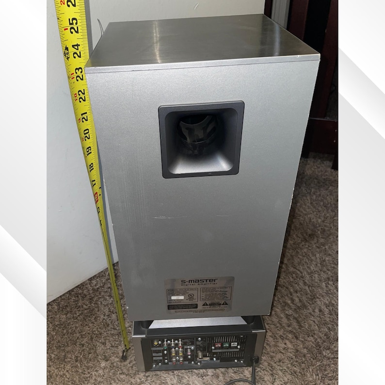 May include: Silver S-master digital audio subwoofer with a black speaker port. The subwoofer is approximately 24 inches tall. The base has multiple input ports. The unit is on a carpeted surface.