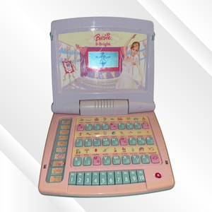 May include: A Barbie B-Bright toy laptop with a lavender screen frame and a pink keyboard base. The screen displays a ballerina graphic and the text Barbie B-Bright. The keyboard features light blue letter keys, pink function keys, and a row of numbered keys.